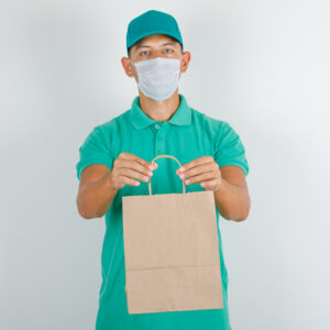 Delivery man holding paper bag in green t-shirt, cap, mask , front view.